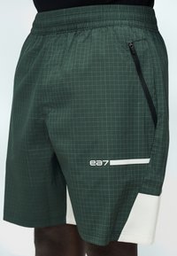 Σκούρα πράσινα καρό σορτς με ελαστική μέση, μαύρη πλαϊνή τσέπη με φερμουάρ, λευκή διχτυωτή επένδυση και λογότυπο "ea7" στο μηρό.