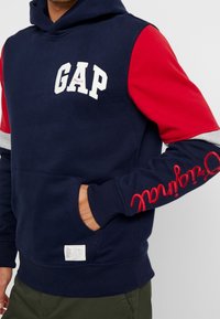 Felpa blu navy con maniche rosse e grigie, con logo "GAP" e dettagli in scrittura. Realizzata in tessuto morbido e con una tasca frontale.