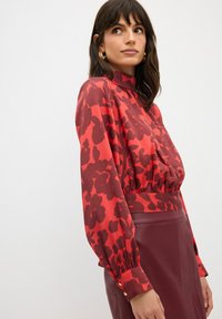 Rode blouse met bloemmotief, gemaakt van lichte stof, met een hoge kraag en lange mouwen, gecombineerd met een bordeauxrode aangesloten leren rok.