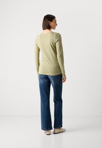 Langermet topp i lys olivenfarge, teksturert stoff; parret med mørkeblå jeans og beige sko. Visning fra baksiden.