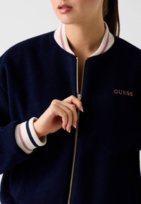 Veste bomber zippée bleu marine avec poignets et col côtelés rayés crème et rose ; présente une fermeture éclair dorée et un petit logo "GUESS" rose.