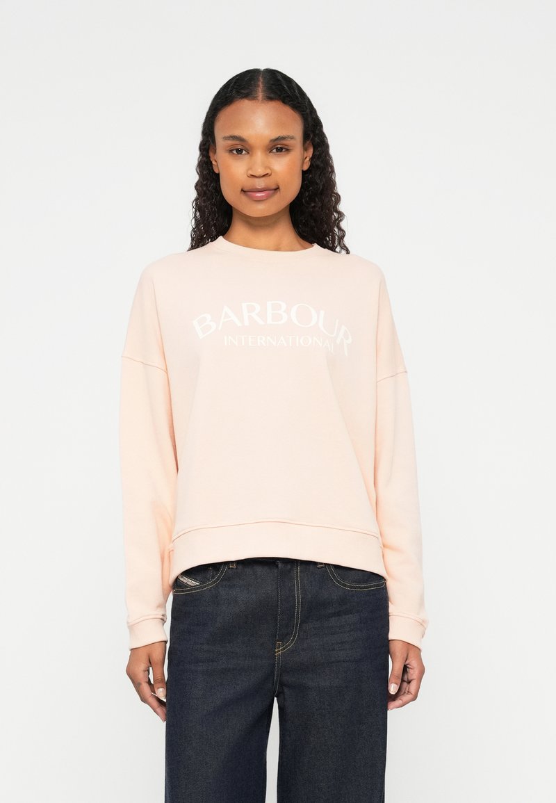 Heled särgis roosa pusa ümmarguse kaeluse, pikkade varrukate ja lõdva istuvusega. Rinnal on valge "BARBOUR INTERNATIONAL" tekst.