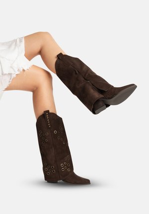 Ben iført mørkebrune ruskind knæhøje støvler med metaløje detaljer, kombineret med hvide, frynset shorts, mod en ensartet baggrund.