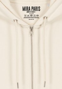 Felpa con zip beige in cotone, caratterizzata da una cerniera argentata, cordini con punte in metallo e etichetta di branding stampata sul colletto.