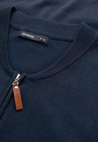 Pull-over zippé marine en tissu doux, avec un col côtelé et une étiquette en cuir marron portant le nom de la marque et la taille.