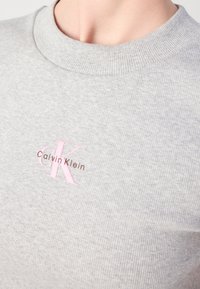 Sivi rebrasti pleteni T-shirt s okruglim izrezom i ružičastim i smešnim Calvin Klein logotipom na prsima.