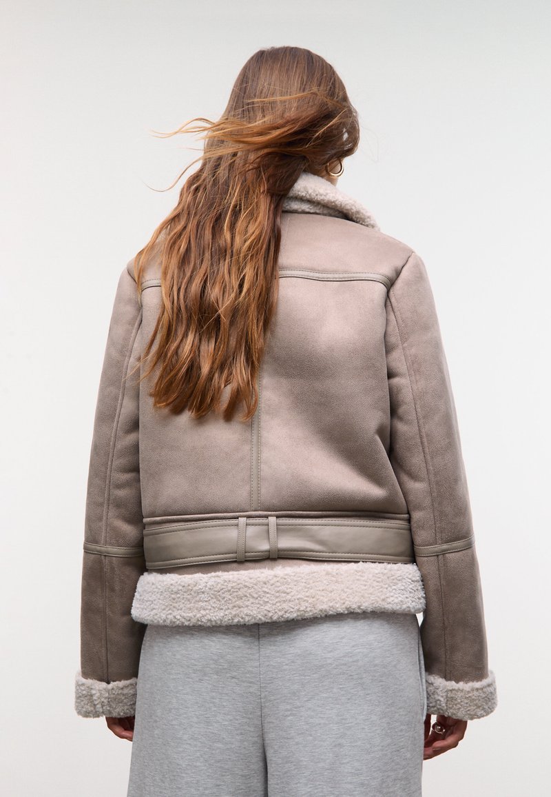Chaqueta de shearling topo con textura suave, corte a la altura de la cintura y un lujoso cuello y dobladillo blanco. Presenta un detalle de cinturón en la cintura.