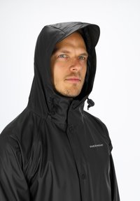 Schwarze wasserdichte Jacke mit hohem Kragen und verstellbarem Kapuze, ausgestattet mit einer durchgehenden Knopfleiste und dezentem Logo-Detail auf der Brust.