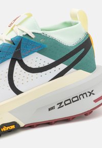 Zapatillas de trail de Nike con una parte superior de malla blanca con acentos en teal, swoosh negro, collar de menta texturizado y una suela intermedia gruesa de color gris con detalles en rojo.