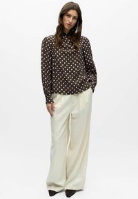 Kvinne står med hendene i lommene på kremfargede bukser med vide ben, iført en brun langermet topp med hvite prikker og svarte sko.
