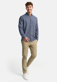 Camisa azul de botones con un patrón texturizado, bolsillo en el pecho, combinada con pantalones beige y zapatillas blancas. El modelo sonríe frente a un fondo claro.