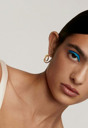 Pendiente de aro dorado con un diseño retorcido y una superficie texturizada. El modelo lleva un delineador azul y un color de labios neutro.