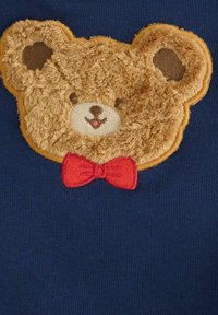 Brązowa twarz misia z puszystą teksturą i ciemnymi uszami, uśmiechająca się, w czerwoną muszkę na granatowym tle.