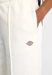 Kreminės spalvos sportinės kelnės, pagamintos iš minkštos medžiagos, su subtiliu logotipo pleistru šone, kišenėmis ir tiesaus kirpimo dizainu.