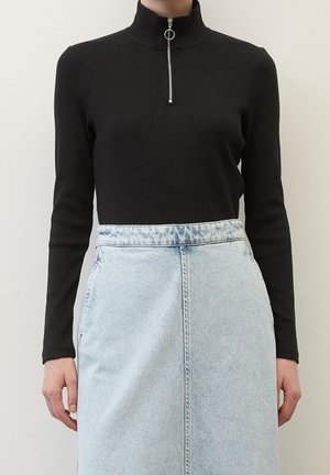 Person trägt ein schwarzes Langarmshirt mit Frontreißverschluss und einen hellblauen, hoch taillierten Jeansrock, steht vor einem schlichten Hintergrund.