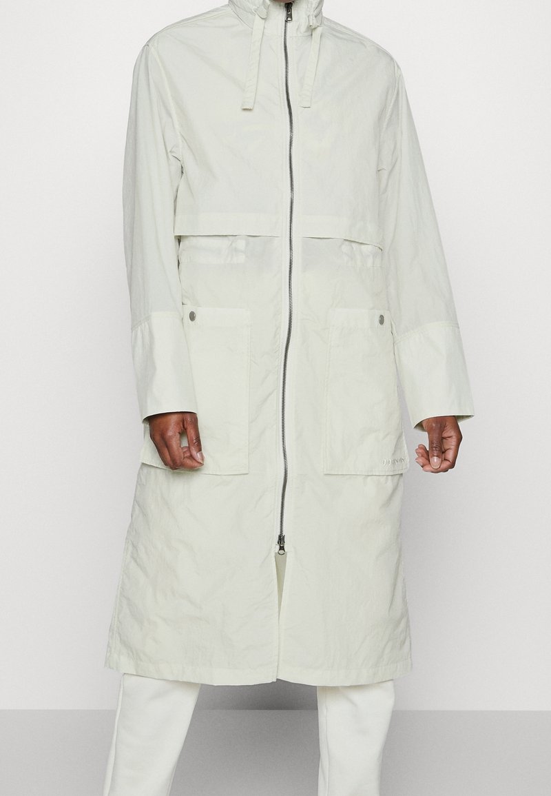 Long manteau beige clair avec fermeture éclair à l'avant, deux grandes poches avant avec boutons et cordons ajustables à la capuche.