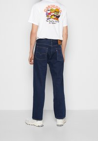 Vit grafisk t-shirt med färggrafisk mattryck på baksidan. Mörkblå denimjeans med klassiska bakfickor och läderlappar. Vita sneakers.