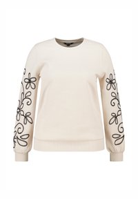 Sweatshirt crème à manches longues avec des broderies florales noires sur chaque manche et des poignets côtelés. Texture lisse avec un col arrondi.