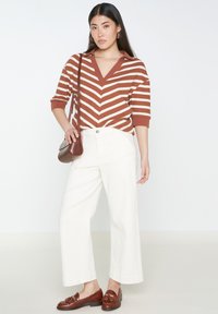 Maglione a righe marroni e crema con scollatura a V, pantaloni bianchi a gamba larga e mocassini marroni. Una piccola borsa a tracolla marrone completa l'outfit.