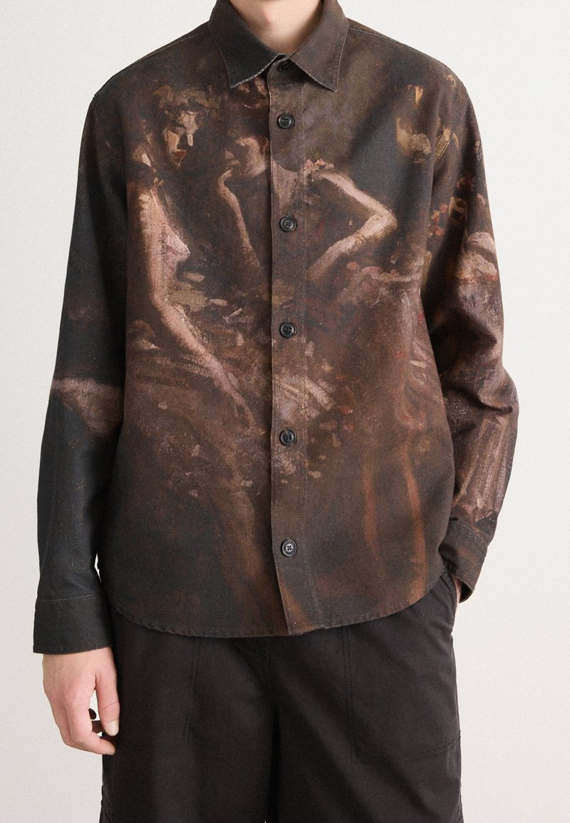 Chemise à manches longues boutonnée avec un imprimé sombre de figures issues d'une peinture classique, portée avec un pantalon noir, montrée des épaules jusqu'aux hanches.