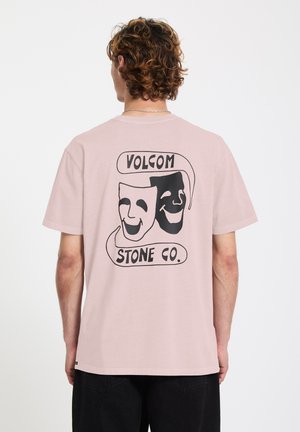 Persoon met krullend haar die een lichtroze t-shirt draagt, met op de achterkant zwarte en witte komedie/tragedie maskers en de tekst "Volcom Stone Co.".