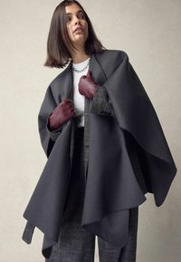 Grey wool cape with oversized design, featuring textured dark grey pants and burgundy leather gloves, layered over a white top.