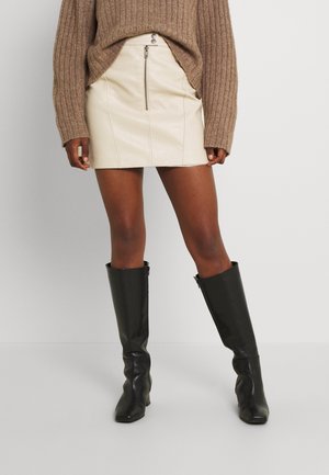 Brown ribbed sweater, cream leather mini skirt with front zipper, and black knee-high leather boots on a standing person.