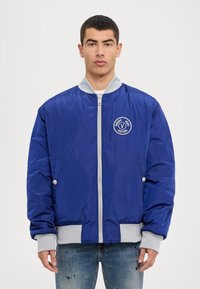 Blue bomber jacket with gray accents, zip closure, ribbed cuffs, and a circular logo on the chest. Smooth texture and slightly oversized fit.
