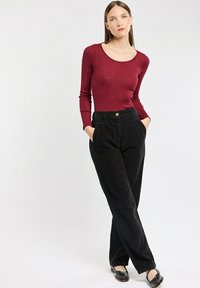 Burgunderfarbiges Langarmoberteil mit Rundhalsausschnitt, kombiniert mit hoch taillierten schwarzen Cordhosen und schwarzen flachen Schuhen.