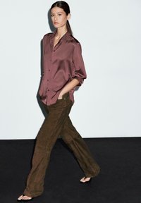 Pantaloni in suede marrone abbinati a una camicia in satin bordeaux con bottoni, caratterizzata da una vestibilità ampia e maniche arrotolate, su uno sfondo minimale.
