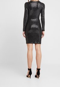 Černé šaty bodycon s dlouhými rukávy a kulatým výstřihem, s těsným střihem a jemnou texturou. Černé vysoké podpatky.