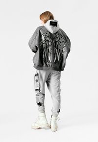 Chaqueta de cuero gris con diseño de alas en la espalda, combinada con pantalones jogger gris claro que presentan gráficos y etiquetas, y botas blancas con cordones.