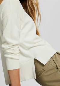 Pull crème à manches longues, poignets côtelés, fentes latérales et coupe décontractée. Associé à un pantalon kaki clair, mettant en valeur un style décontracté.