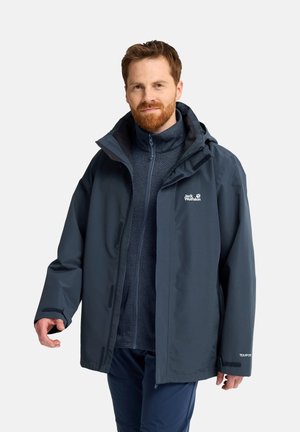 Mann trägt dunkelblaue Outdoorjacke und Reißverschlusspullover, steht vor hellem einfarbigem Hintergrund, schaut nach vorne mit leichtem Lächeln.