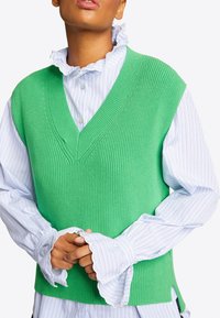 Person trägt einen leuchtend grünen gestrickten ärmellosen Pullover über einer weißen Bluse mit blauen Streifen und Rüschenkragen sowie -ärmeln.