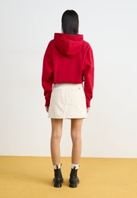 Una donna sta in piedi di spalle indossando un maglione rosso corto, una gonna beige chiaro a righe, calzini bianchi e stivaletti neri su un pavimento senape.