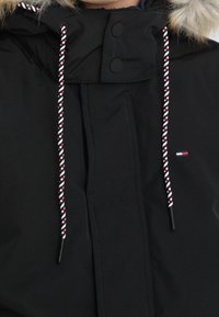 Detailní záběr černé zimní bundy s kapucí lemovanou kožešinou, pruhovanými šňůrkami, zapínáním na knoflíky a malým vlajkovým logem na hrudi.