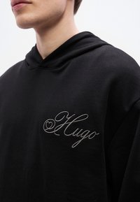 Μαύρο φούτερ με κουκούλα από μαλακό ύφασμα, με τσεπάκι μπροστά. Γκρι κεντημένο λογότυπο "Hugo" με διακοσμητική καρδιά στην αριστερή πλευρά του στήθους.
