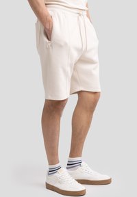 Lys beige sportshorts lavet af tekstureret stof, med sidelommer og snørebånd i taljen, parret med hvide sneakers og stribede sokker.