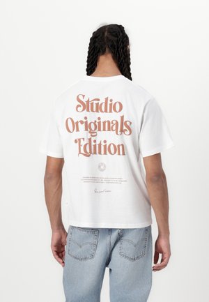 Witte t-shirt met korte mouwen, voorzien van grote, retro-stijl oranje tekst op de achterkant: "Studio Originals Edition." Zacht katoen materiaal.
