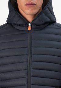 Gevoerde marineblauwe jas met een ritssluiting aan de voorkant en een capuchon. Voorzien van horizontale stiksels en een contrasterende oranje ritslip voor extra detail.