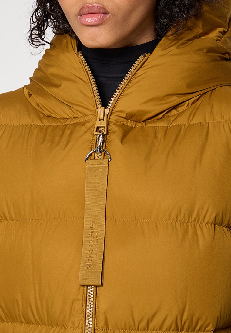 Musztardowa żółta kurtka ocieplana z wysokim kołnierzem, zamkiem błyskawicznym z przodu i wytłoczonym logo na metce. Charakteryzuje się gładką teksturą oraz ciepłym designem.