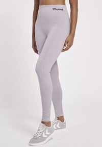 Licht lavendelkleurige high-waisted legging met gestructureerde lijnen en een klein zwart logo op de tailleband, gecombineerd met grijze sneakers met witte accenten.