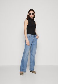 Black sleeveless mock-neck top, light blue wide-leg jeans, and light brown pointed-toe flats with hardware detail. Model wearing oversized sunglasses.