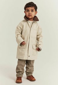 Beige vattentät kappa med brun krage, två frampåsar, bärs över en mönstrad skjorta och kombineras med ljusa khakibukser och bruna skor.