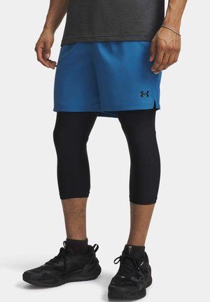 Mann trägt blaue Sportshorts über schwarzen kurzen Leggings und schwarzen Sportschuhen, steht vor einem schlichten Hintergrund.