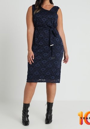 Marineblauwe kanten jurk met een aansluitend silhouet, een V-hals en een riem in de taille. Voorzien van bloemmotieven en een mouwloze stijl, gecombineerd met zwarte laarzen.