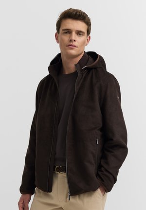 Junger Mann, der eine dunkelbraune Zip-up-Jacke mit Kapuze, ein schwarzes Shirt, beige Hosen trägt und mit den Händen in den Taschen vor einem einfarbigen hellen Hintergrund steht.