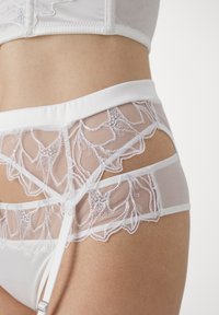 Balta apatinė lingerie, pasižyminti permatomu tinkleliu su gėlių nėriniu, reguliuojamomis petnešomis ir lygiu juosmeniu. Elegantiškai ir sudėtingai sukurta.