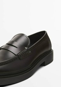 Black leather loafers with a braided design across the upper, featuring a low block heel and smooth texture. Stitched detailing is visible.